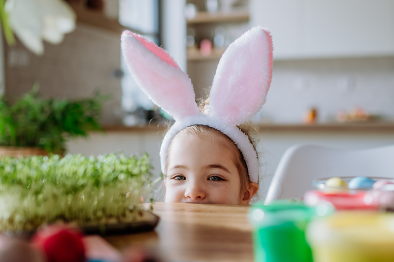 Wielkanocne godziny otwarcia w M1 Częstochowa. Dziewczynka w uszach zająca wielkanocnego, wychylająca się zza stołu.