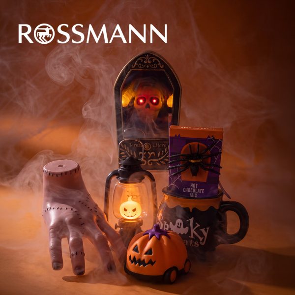 Dekoracje na Halloween na pomarańczowym tle spowitym dymem: sztuczna ręka ze szwami, dzwonek z czaszką o świecących czerwonych oczach, latarnia z podświetlaną dynią, pojazd w kształcie dyni oraz kubek z opakowaniem czekolady do picia ozdobionym pająkiem. U góry białe logo Rossmann.